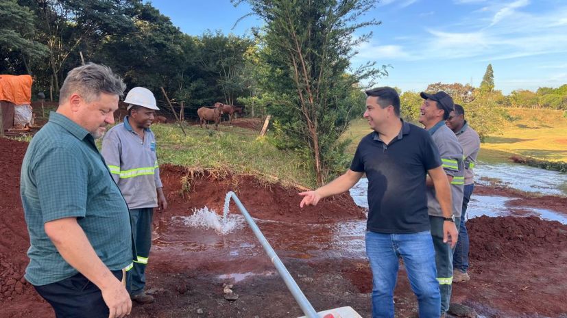 Água jorrando da tubulação do novo poço, com a equipe do SAAE reunida ao redor da perfuração.
